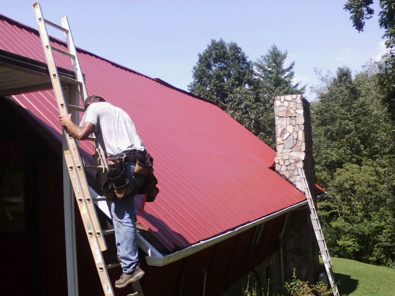 Roofer on a ladder beside a red metal roof and stone chimney — Soffit & Fascia Repair by Roofing Brothers in Northvale